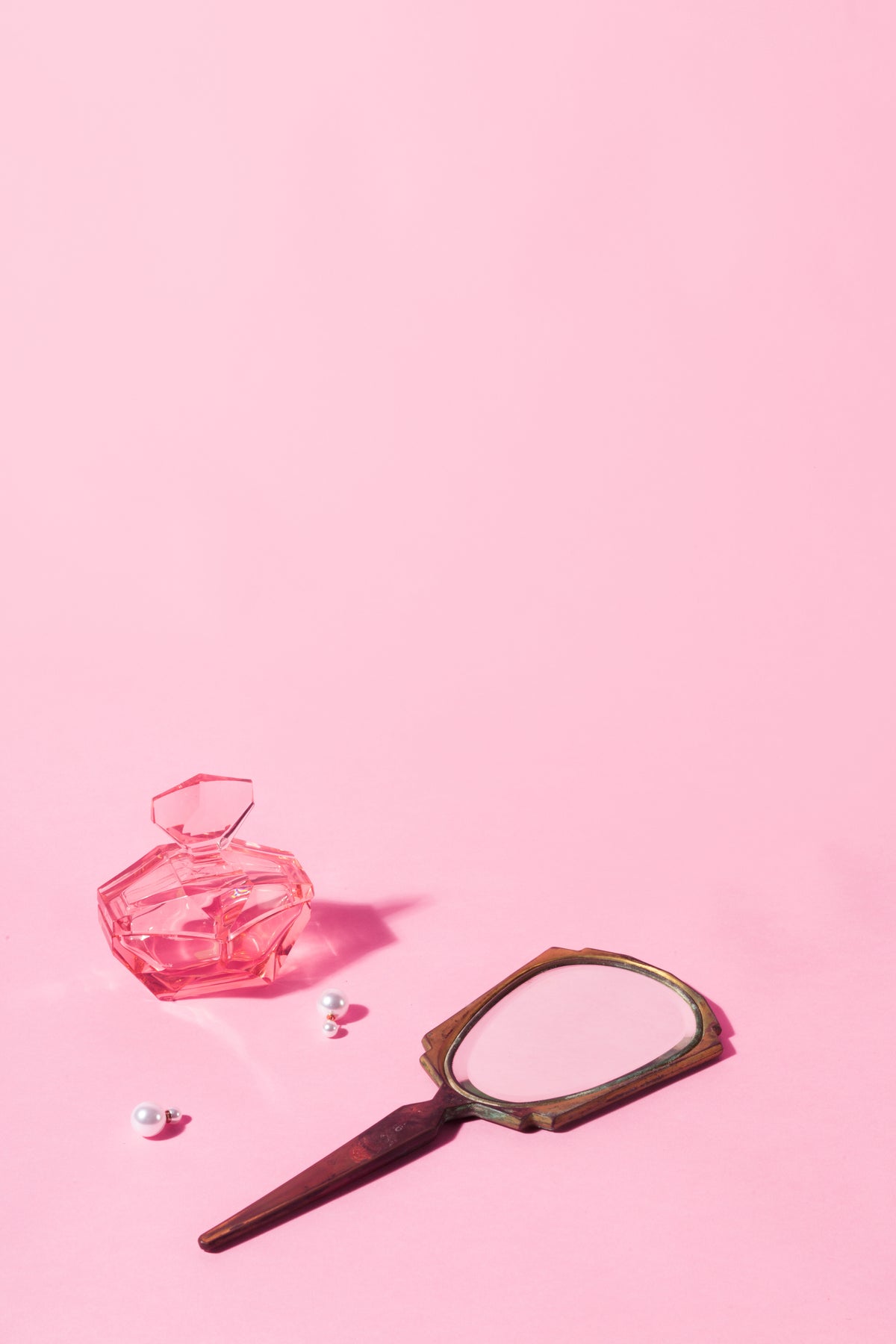 A handheld mirror, perfume bottle, and earrings on a pink surface.