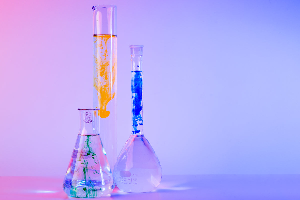 Glass laboratory vessels containing colored liquids against a pastel background.