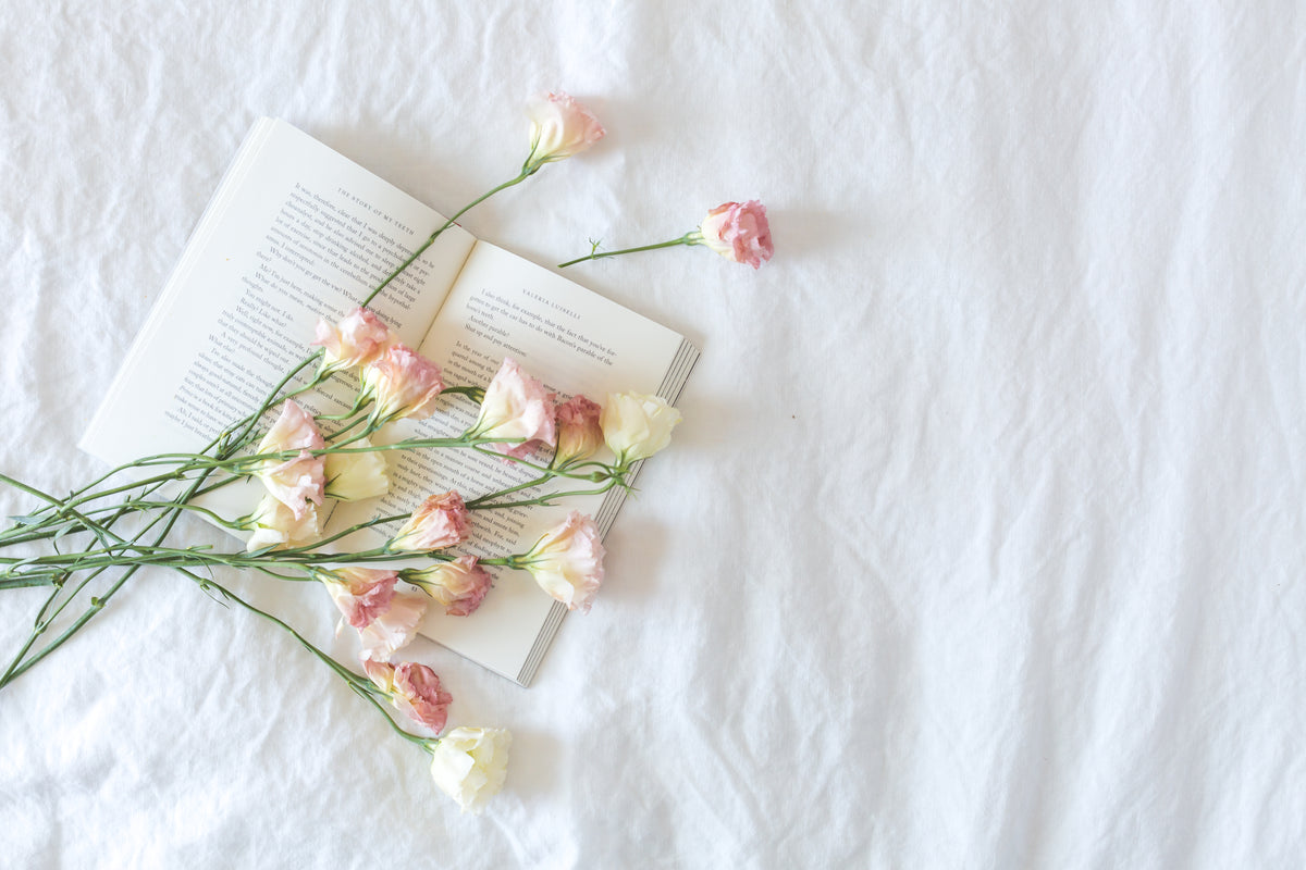 An open book and flowers rest on white bedding.