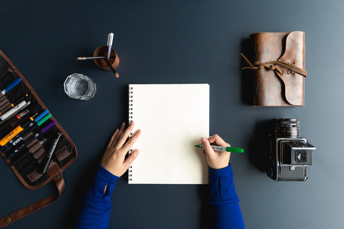 Overhead view of a person writing in a notebook beside a camera and pencil case.