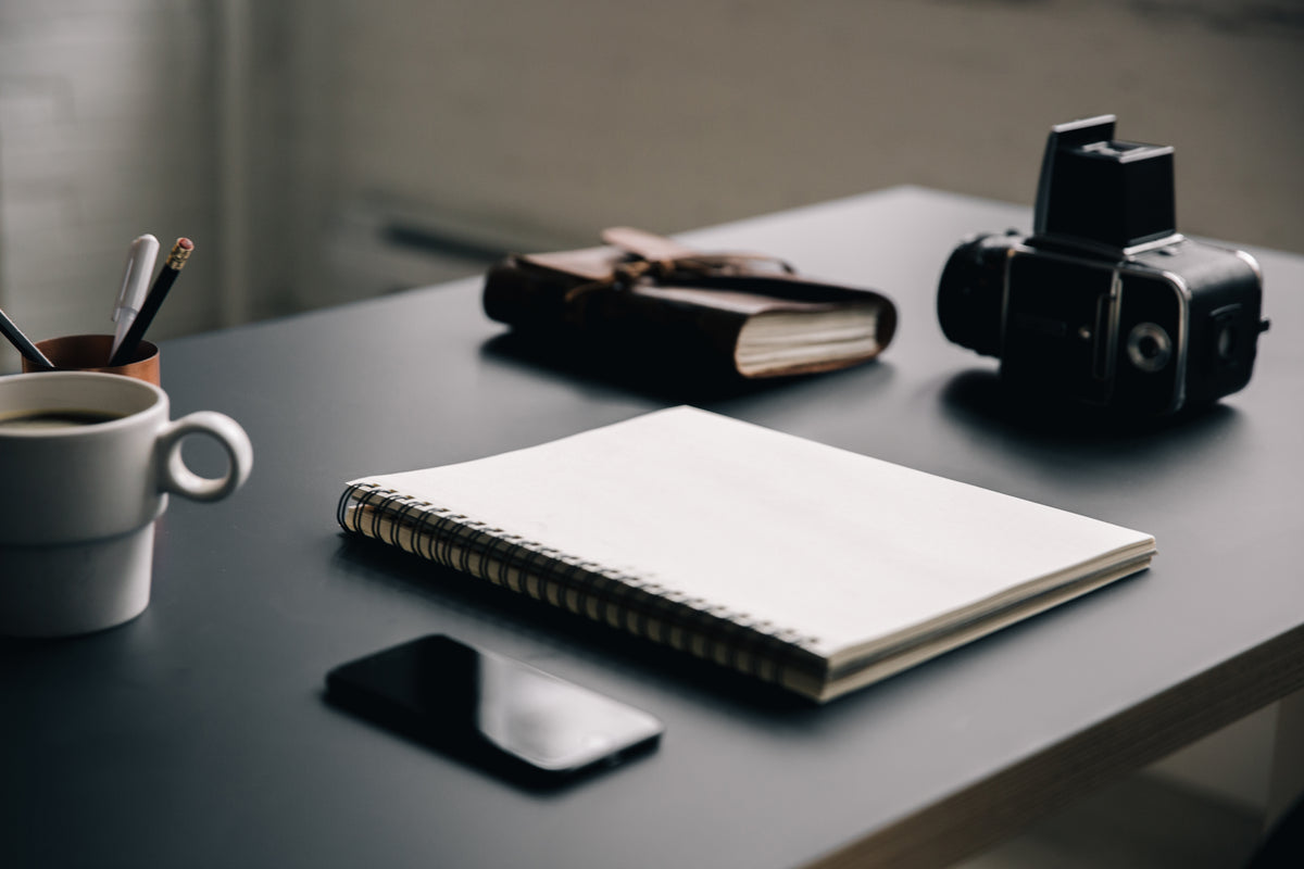 A blank notebook lies on a desk beside a mug and camera.