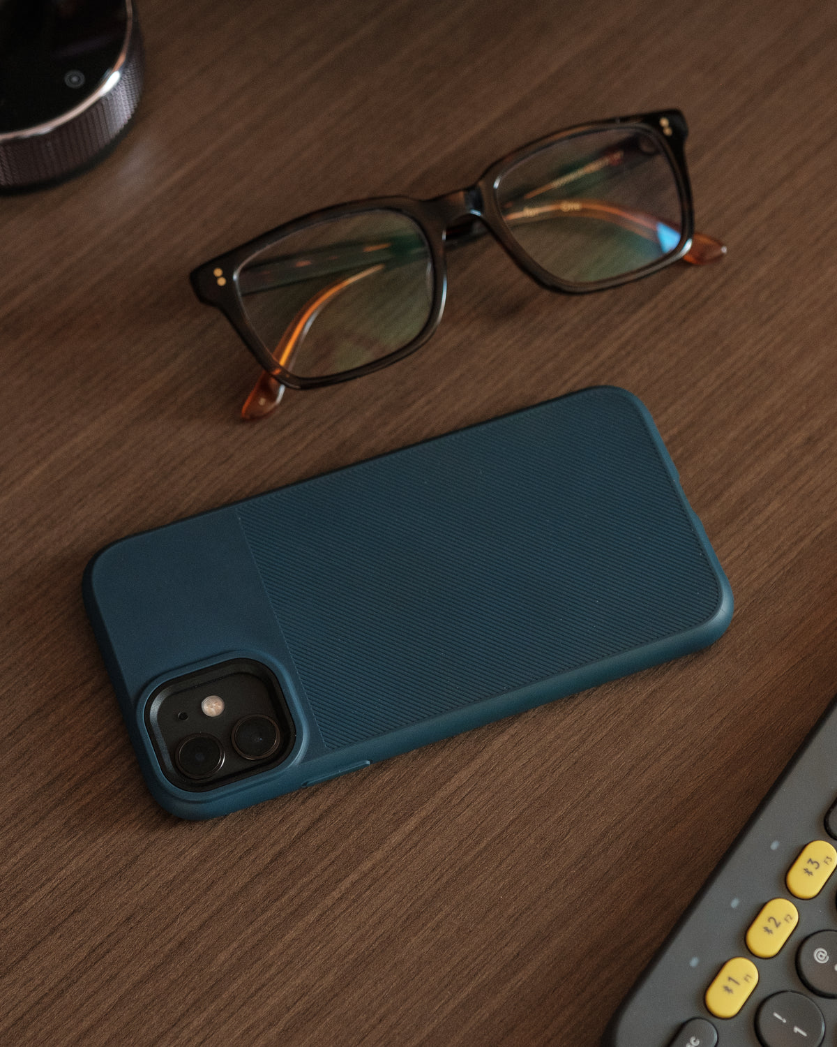 A phone in a blue case and eyeglasses rest on a wooden table.