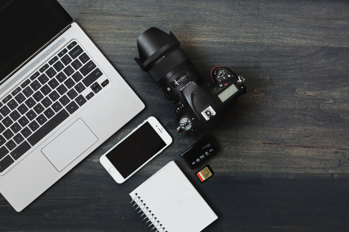 A camera, phone, laptop, and notebook lie on a dark tabletop.