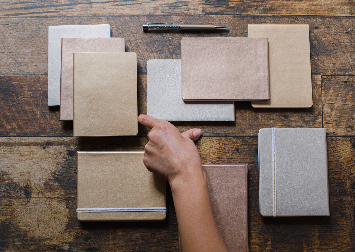 Notebooks and pens lie on a wooden table beside a person’s hand.