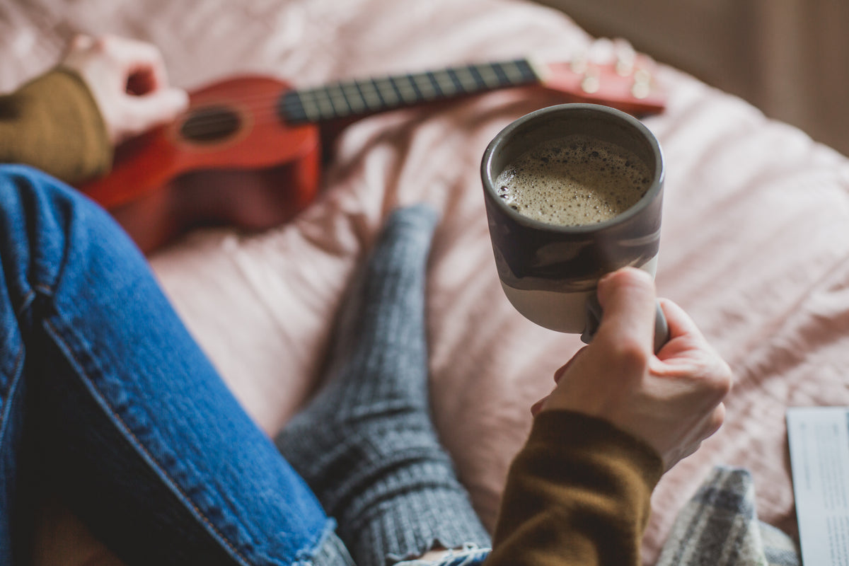 A person holds a cup of coffee while sitting on a bed beside a small guitar.