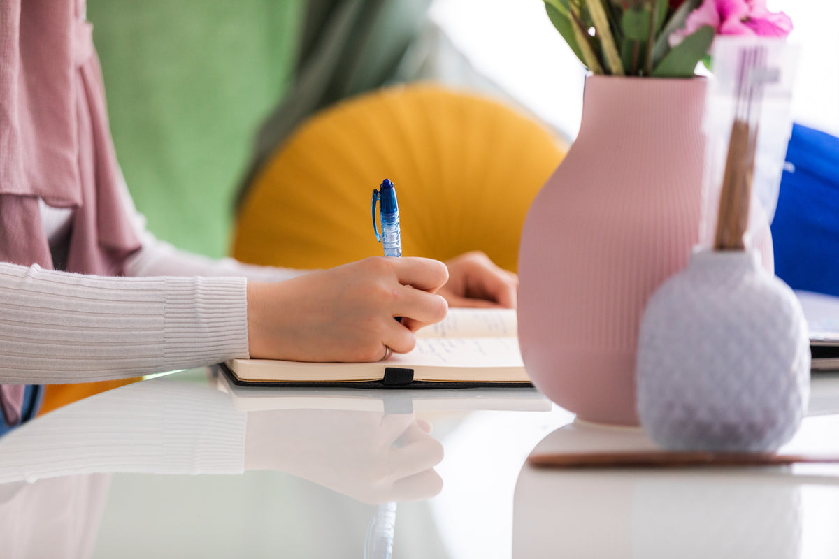 A hand writes notes in a planner at a colorful workspace.