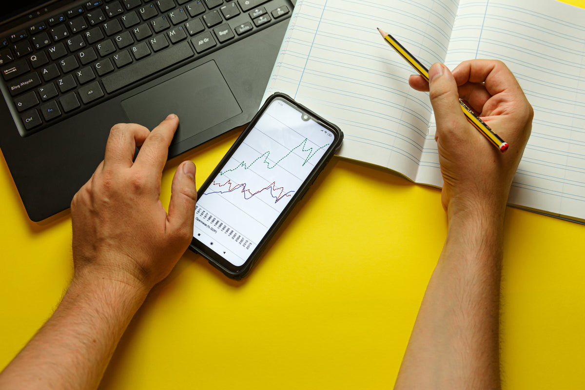 Hands take notes beside a laptop and smartphone on a yellow desk.