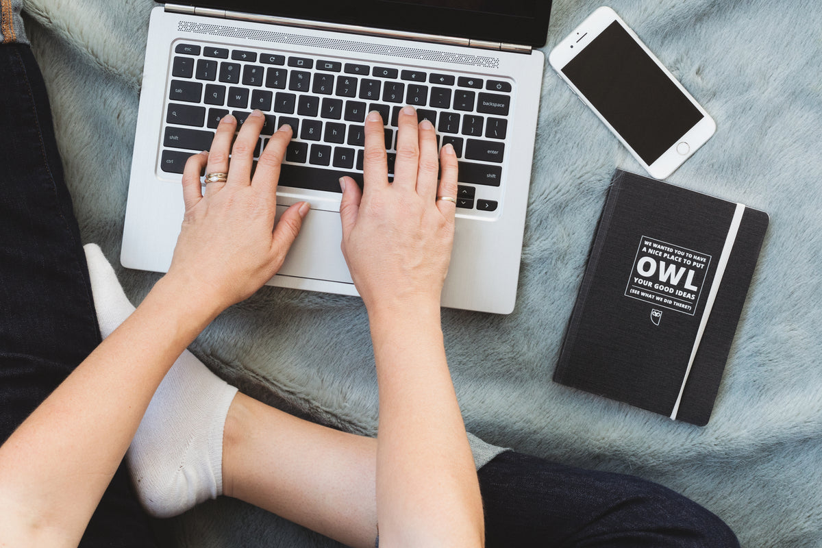 A person works on a laptop with a phone and notebook nearby.
