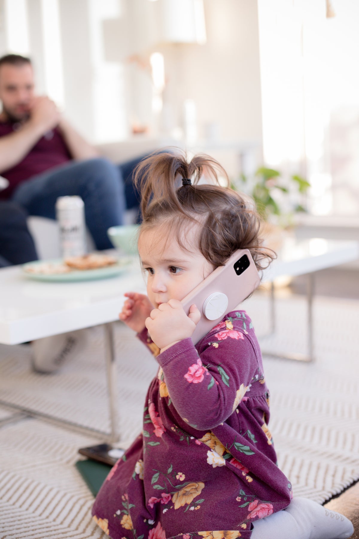 A child holds a pink smartphone while an adult sits in the background.