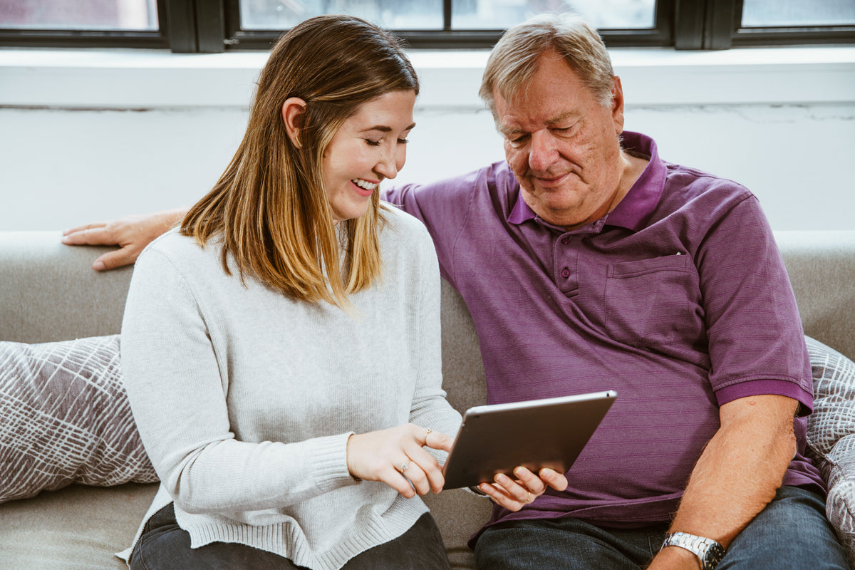 A younger adult sits beside an older adult while they use a tablet together.