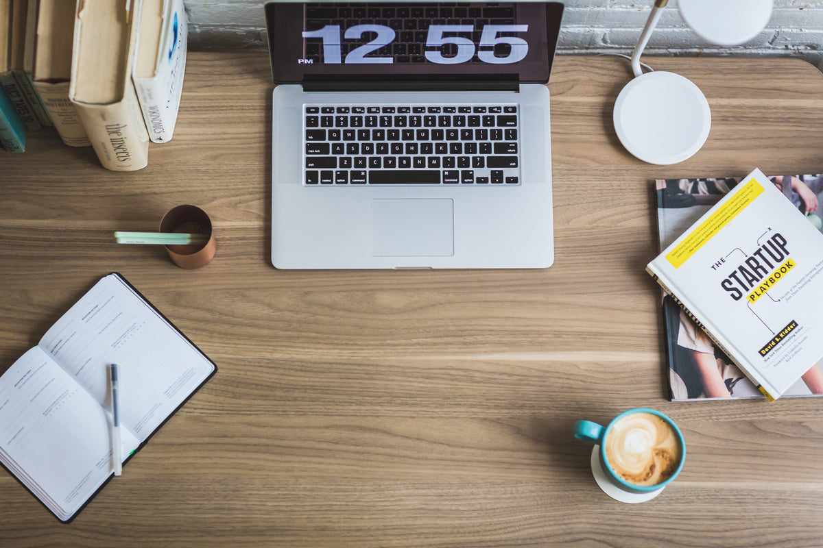 A laptop showing a clock sits on a wooden desk beside a notebook.