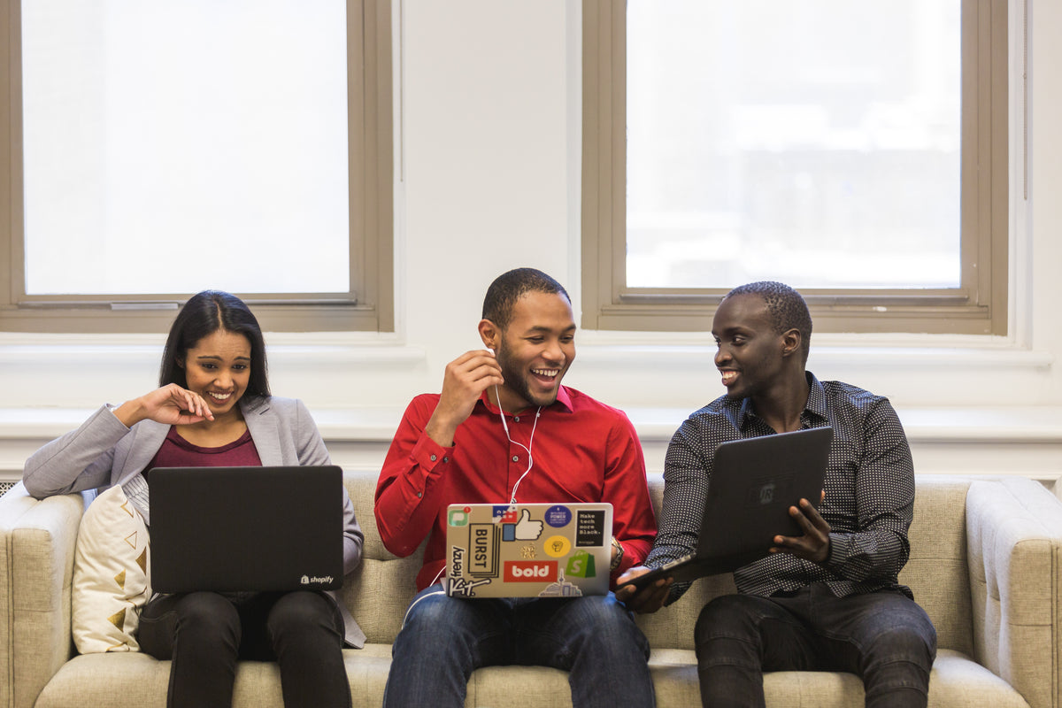 Three people smile while using laptops on a couch.