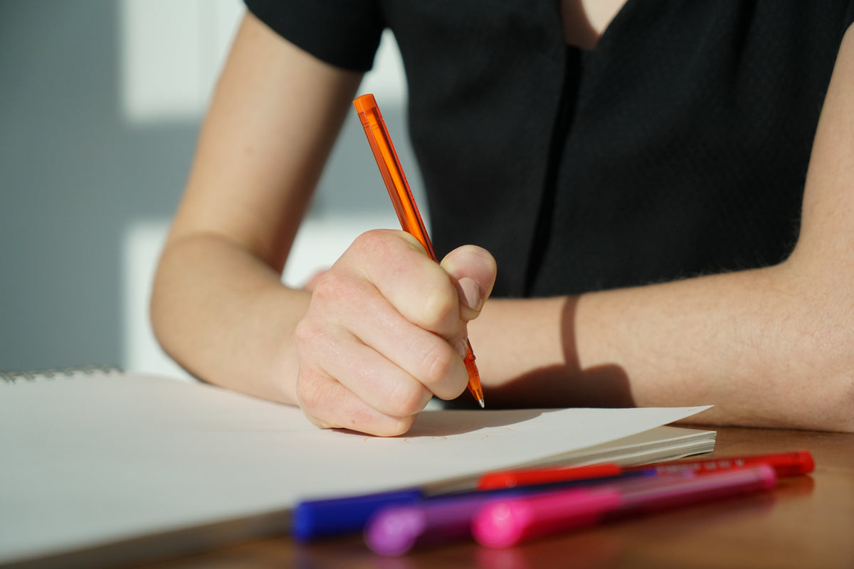 A person writes with a pencil on paper beside colored pens.