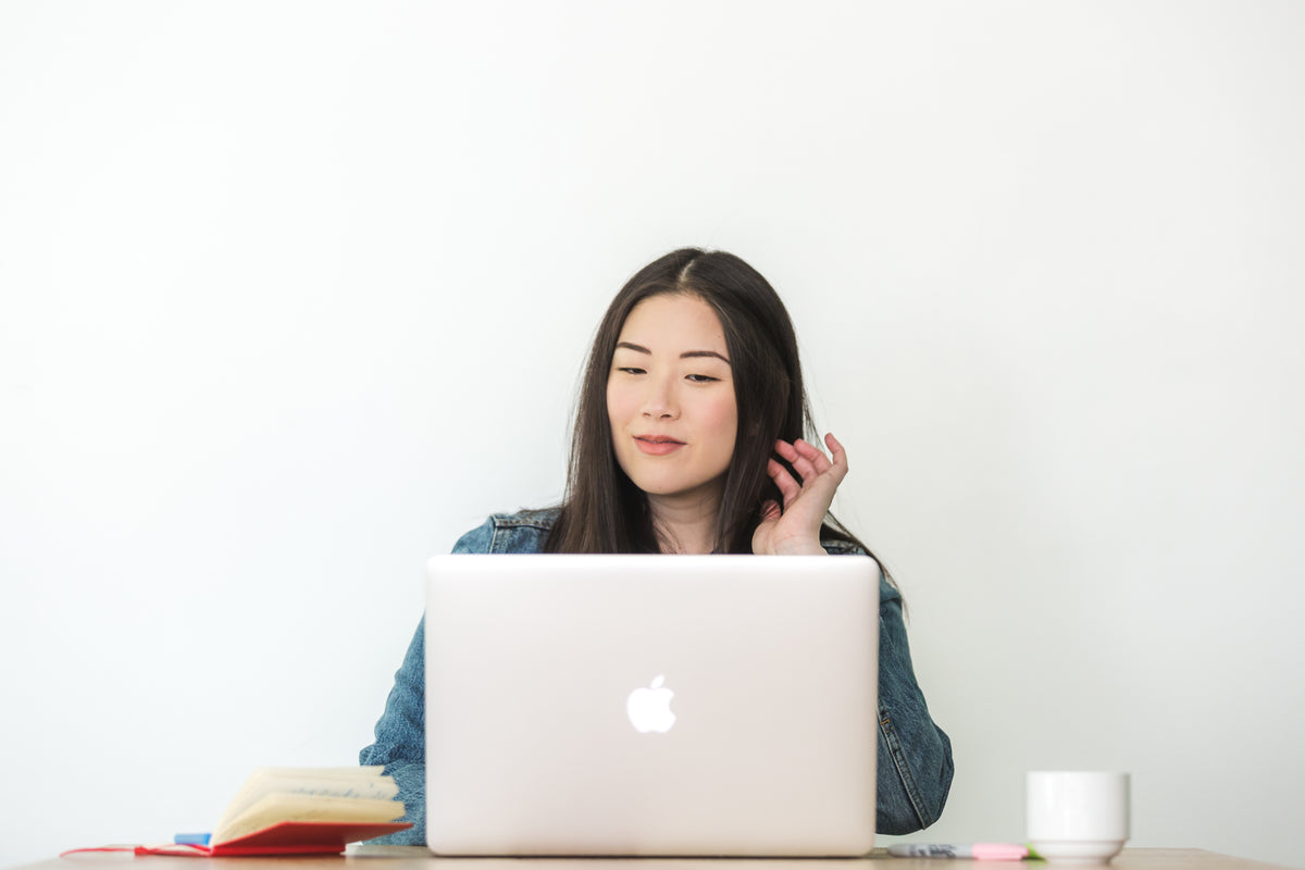 A woman sits behind a laptop with a notebook nearby.