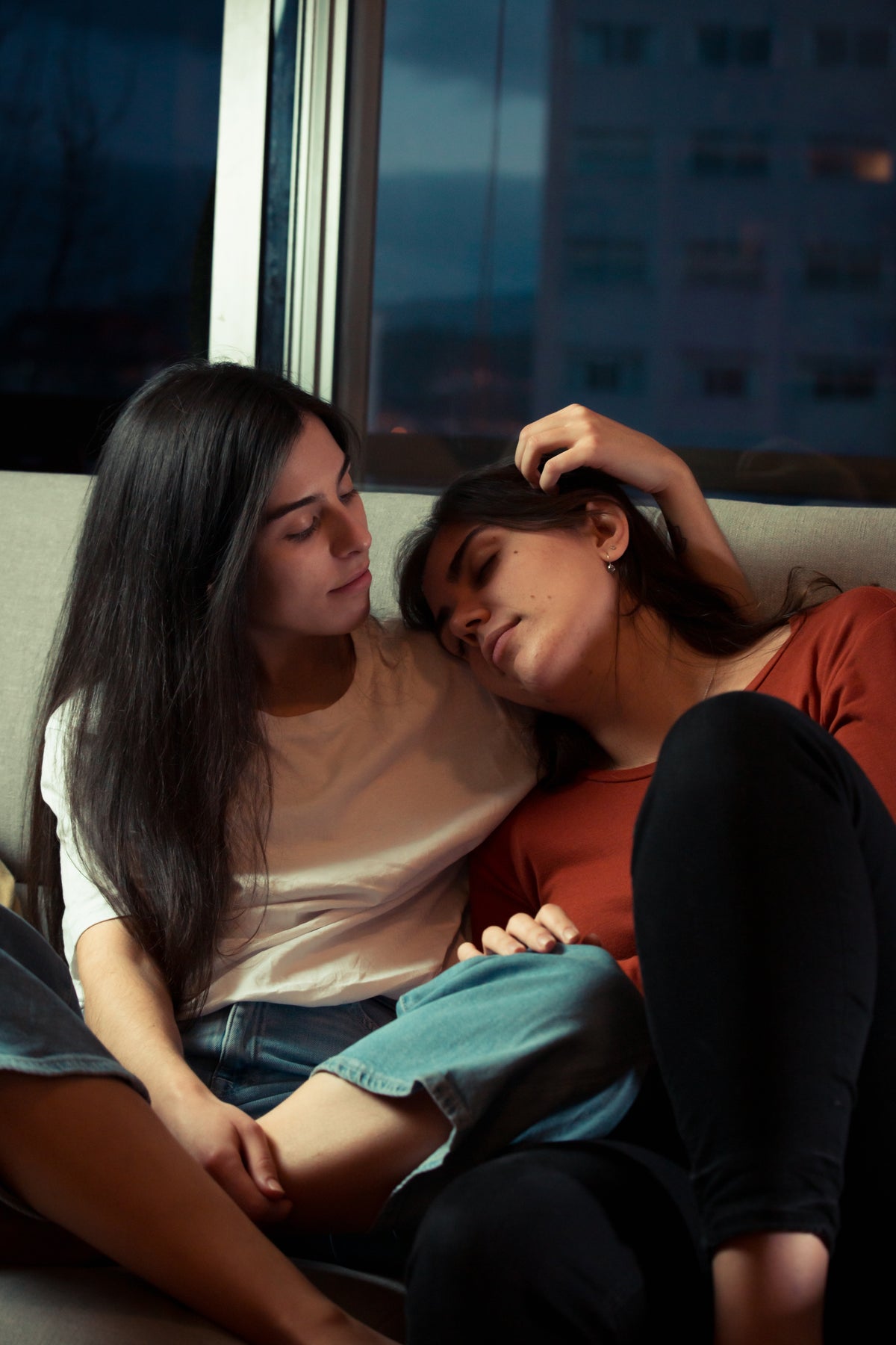 Two people sit close together in a quiet living room, one resting her head on the other's shoulder.