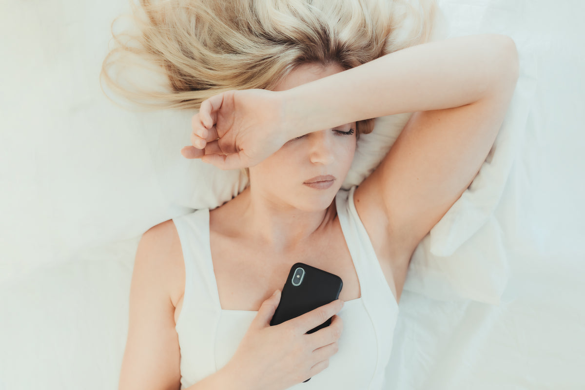 A woman lying in bed holds a phone with one arm across her face.