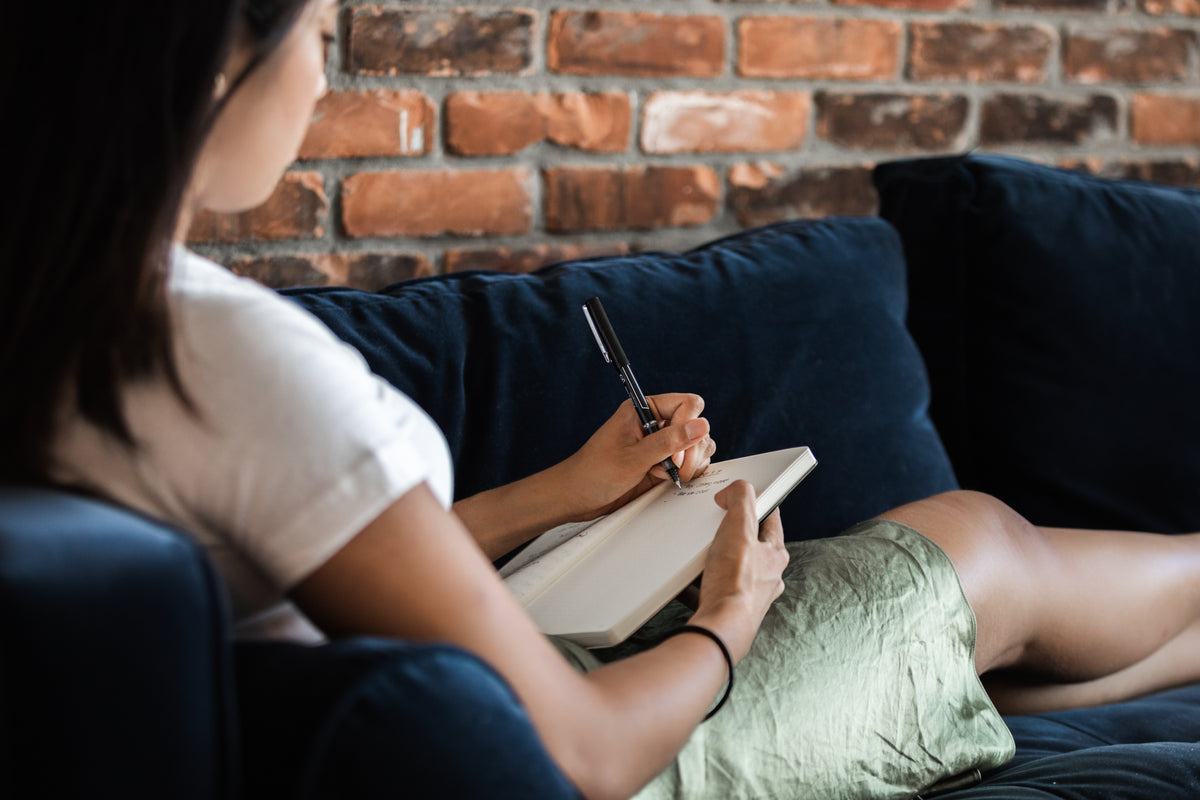 A person writes in a notebook while seated on a sofa.