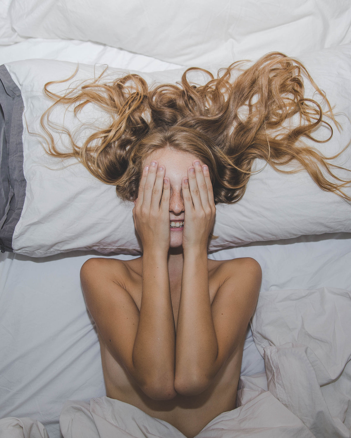 A woman lies on white bedding with her hands over her eyes.
